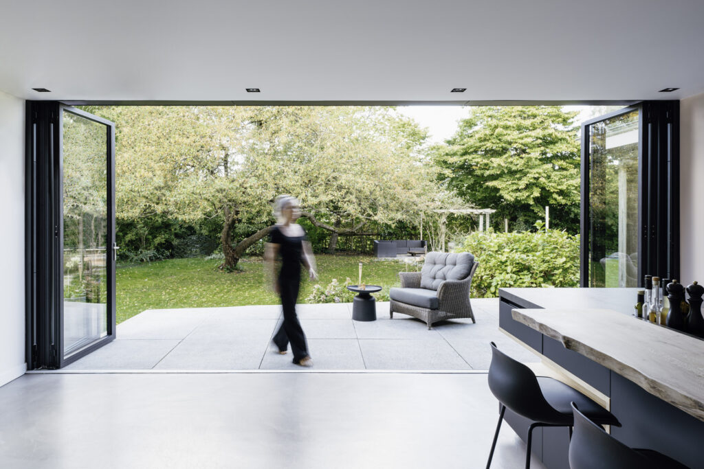Moderne Wohnküche mit Blick auf einen Garten, in dem eine Person mit grauem Haar vorbeigeht. Die großen Fenster öffnen sich zur Terrasse, die mit einem gemütlichen Sessel und einem kleinen Tisch ausgestattet ist. Herbstliche Bäume im Hintergrund schaffen eine einladende Atmosphäre.