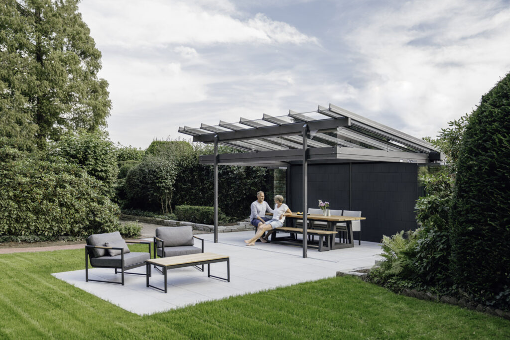 Paar sitzt entspannt unter einer modernen Überdachung auf einer Terrasse, umgeben von grünem Garten. Es gibt eine Essgruppe und gemütliche Sitzgelegenheiten.