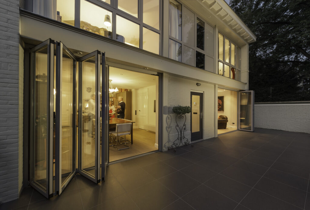 Moderne Architektur eines Hauses mit großen Fenstern, offenen Falttüren und einem einladenden Innenraum, der bei Nacht beleuchtet ist. Die Terrasse ist mit dunklen Fliesen ausgestattet und bietet einen Blick auf die gemütliche Atmosphäre im Inneren, wo sich Personen um einen Esstisch versammeln.