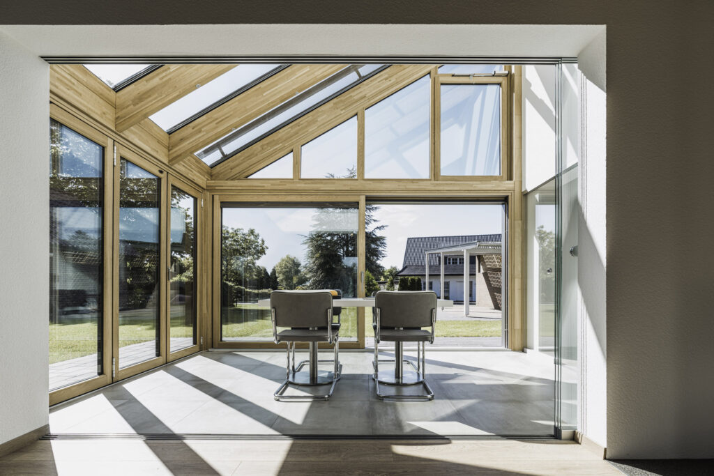 Helles Wintergarten-Design mit großen Fenstern und Holzrahmen, ausgestattet mit zwei modernen Stühlen und einem Tisch, Blick auf einen gepflegten Garten und ein Haus im Hintergrund.