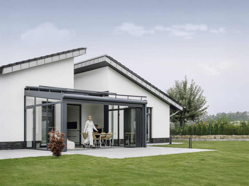 Moderne Wohnarchitektur mit einem eleganten Anbau und großzügigen Glasfronten, die einen Blick auf den Garten bieten. Eine Frau verlässt das Haus und genießt den Außenbereich mit grünem Rasen und einem gepflegten Garten.
